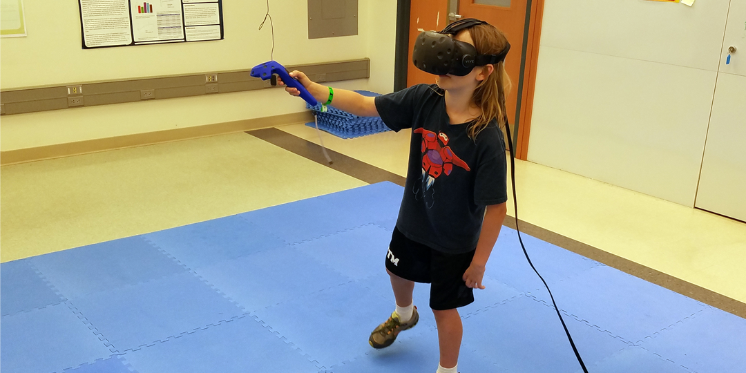 A young child tosses an HTC Vive controller to an experimenter who is posed to catch it. An inflatible air mattress and tiled foam squares are on the ground to protect the equipment from errant tosses.