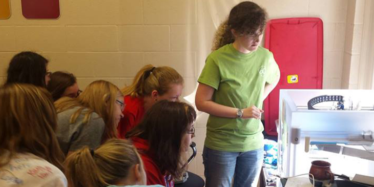 Female middle and high school students crowd around a 3D printer, which is supervised by an undergraduate camp counselor. 