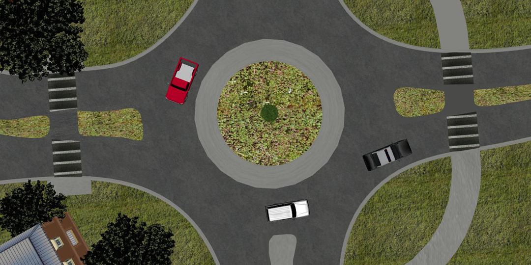 A birds-eye view of the virtual environment is presented. A roundabout is displayed in which three cars are circulating. Sparse trees and buildings populate the area around the traffic circle 